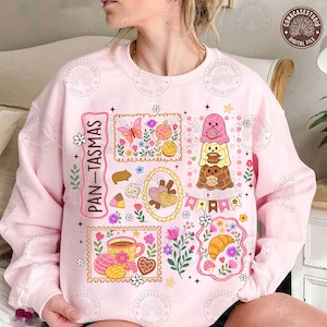 May include: A light pink sweatshirt featuring a colorful graphic print with the text PAN-TASMAS. The design includes illustrations of various breads, pastries, flowers, a butterfly, and three stacked ghost characters, all arranged in decorative frames.