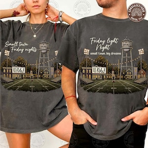 Puede incluir: Una camiseta gris carbón con un gráfico de un campo de fútbol americano de noche, una torre de agua, luces de estadio y el texto "Small town Friday nights" o "Friday light Night small town big dreams" con el número 47567 en el marcador.