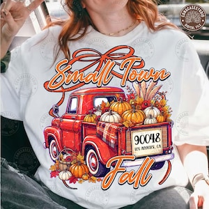 May include: A white t-shirt featuring a graphic of a red pickup truck filled with pumpkins and autumn foliage. The text reads Small Town Fall and 90048 Los Angeles, CA. The design includes a large orange bow above the truck and a plaid blanket.
