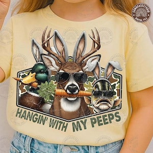 May include: A light yellow t-shirt featuring a graphic of a deer, duck, and fish wearing sunglasses and bunny ears, each holding a carrot. The text "HANGIN' WITH MY PEEPS" is below the graphic, set against a camouflage background.