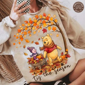 May include: A beige sweatshirt featuring a graphic of Winnie the Pooh and Piglet in an autumn forest. Pooh wears a red shirt and scarf, while Piglet sits in a wooden wagon filled with pumpkins and leaves. The text reads 'tis the season'.