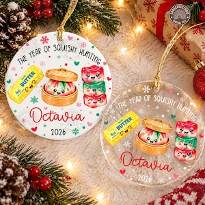 以下が含まれることがあります： 「THE YEAR OF SQUISHY HUNTING」、「Octavia」、「2026」の文字が入った2つの丸いクリスマスオーナメント。黄色い有塩バター、竹製の蒸し器に入った点心、積み重なった3つのカラフルなスクイーズ玩具のイラストが描かれています。
