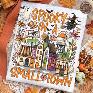 Könnte beinhalten: Eine weiße rechteckige Fläche mit einer farbenfrohen Halloween-Illustration und dem Text SPOOKY IN A SMALL TOWN. Das Design zeigt einen Geist, eine Hexe auf einem Besen, einen Wasserturm und eine Häuserreihe mit orangefarbenen und violetten Akzenten.