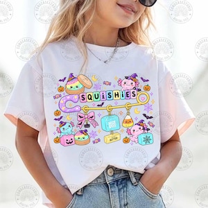 May include: A white t-shirt featuring a graphic print of a large yellow safety pin with the text SQUISHIES. The design includes pink axolotls, colorful squishy toys, pumpkins, bats, spiderwebs, and a pink bow, all arranged around the safety pin.