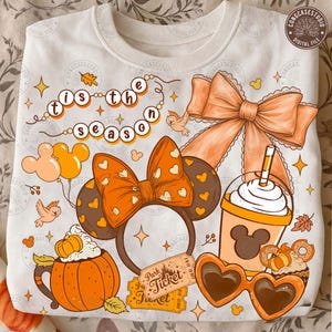 May include: A white sweatshirt featuring a graphic print with the text 'tis the season', orange mouse ears with hearts, a large orange bow, a pumpkin-shaped mug, a drink with a mouse-shaped logo, heart-shaped sunglasses, and a park ticket.