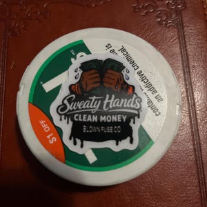 May include: A white, round container with a green and white rim. The container has a sticker with the text "Sweaty Hands Clean Money Blown Fuse Co" and a graphic of hands holding money. A sticker reads "$1 OFF".