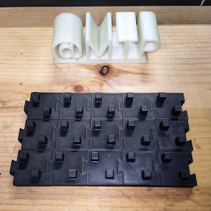 Op de afbeelding: Twee 3D-geprinte objecten op een houten oppervlak. Eén is een lichtgroen "@MTP"-ontwerp. De andere is een zwart, in elkaar grijpend tegelsysteem met vierkante uitsteeksels. Waarschijnlijk prototypes of componenten.