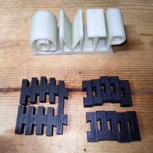 May include: A 3D-printed object with the letters "OMP" in white, and several black, interlocking pieces. The letters are on a base, and the interlocking pieces are arranged below. The objects are on a wooden surface.