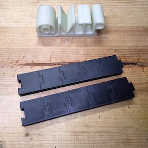 May include: Two black, interlocking, rectangular pieces and a white 3D-printed object spelling "OMTP" on a wooden surface. The "O" is a cylinder, and the "M" and "P" are rectangular.