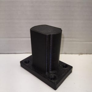 May include: A black, 3D-printed bracket with a rectangular base and a vertical, curved top section. The base has four holes for mounting. The object is made of a solid, matte black material and sits on a white surface.