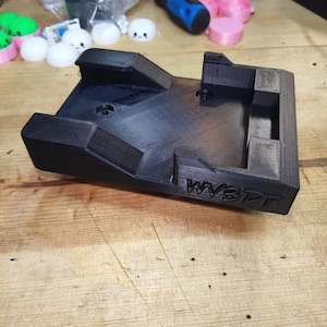 May include: A black 3D printed rectangular mounting bracket with a recessed center, raised side walls, and two circular screw holes. The front edge features the embossed text WV3DT. The object has a textured surface finish from the printing process.