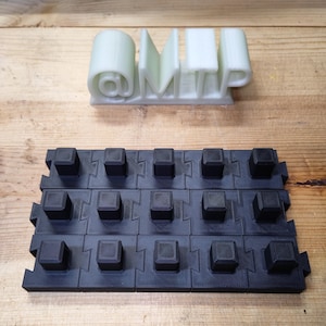 Puede incluir: Vista superior de dos objetos impresos en 3D. El primero es un texto verde claro que dice "@MTP". Debajo, se ve un sistema de baldosas negras entrelazadas. Las baldosas tienen protuberancias cuadradas.