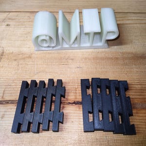 May include: A light green 3D-printed sign with the letters "@MTP" and a rolled shape. Below are two black, geometric, interlocking shapes. The objects are on a wooden surface.