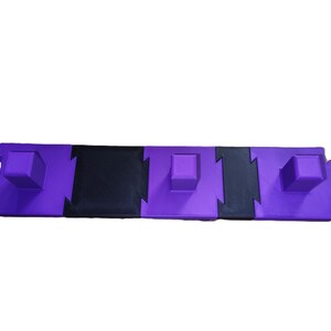 May include: A modular purple and black foam play mat. The mat is composed of interlocking pieces, with purple squares and black rectangular sections. The design includes raised square blocks on the purple sections, creating a textured surface.