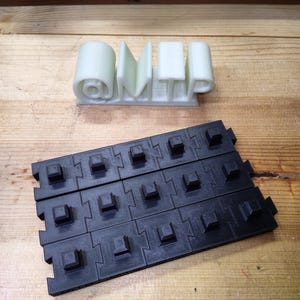 Könnte beinhalten: Ein hellgrünes 3D-gedrucktes Schild mit der Aufschrift "@MTP" in einer stilisierten Schriftart. Darunter befindet sich ein schwarzes, ineinandergreifendes, modulares Fliesensystem mit quadratischen Vorsprüngen. Die Objekte befinden sich auf einer Holzoberfläche.