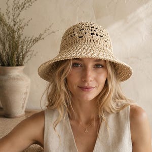 May include: A beige, open-weave sun hat with a wide brim. The hat has a rounded crown and a slightly flared brim. The hat is made of a natural fiber, likely straw or raffia. The hat is worn by a person wearing a beige sleeveless top.