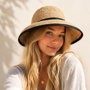 May include: A beige sun hat with a black band and trim. The hat has a wide brim and a rounded crown. The hat is made of woven material. The hat is worn by a person with long blonde hair.