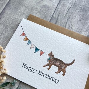 May include: A white greeting card with a textured surface featuring a watercolor illustration of a brown tabby cat holding a string of colorful triangular bunting flags in its mouth. The text Happy Birthday is printed in black below the cat.