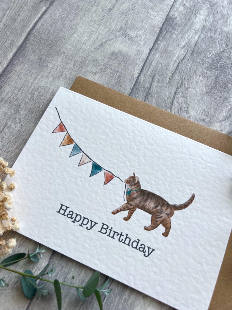 May include: A white greeting card with a textured surface featuring a watercolor illustration of a brown tabby cat holding a string of colorful triangular bunting flags in its mouth. The text Happy Birthday is printed in black below the cat.