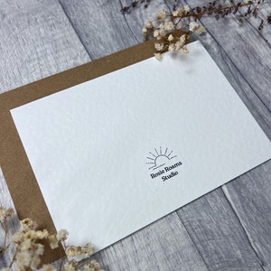 May include: A white card with a brown envelope behind it. The card has a simple black and white logo with a sun rising over waves and the text "Rosie Roams Studio".