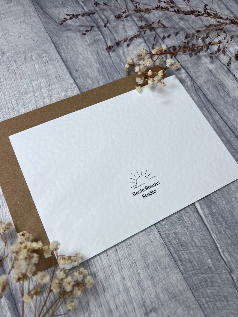 May include: A white card with a brown envelope behind it. The card has a simple black and white logo with a sun rising over waves and the text "Rosie Roams Studio".