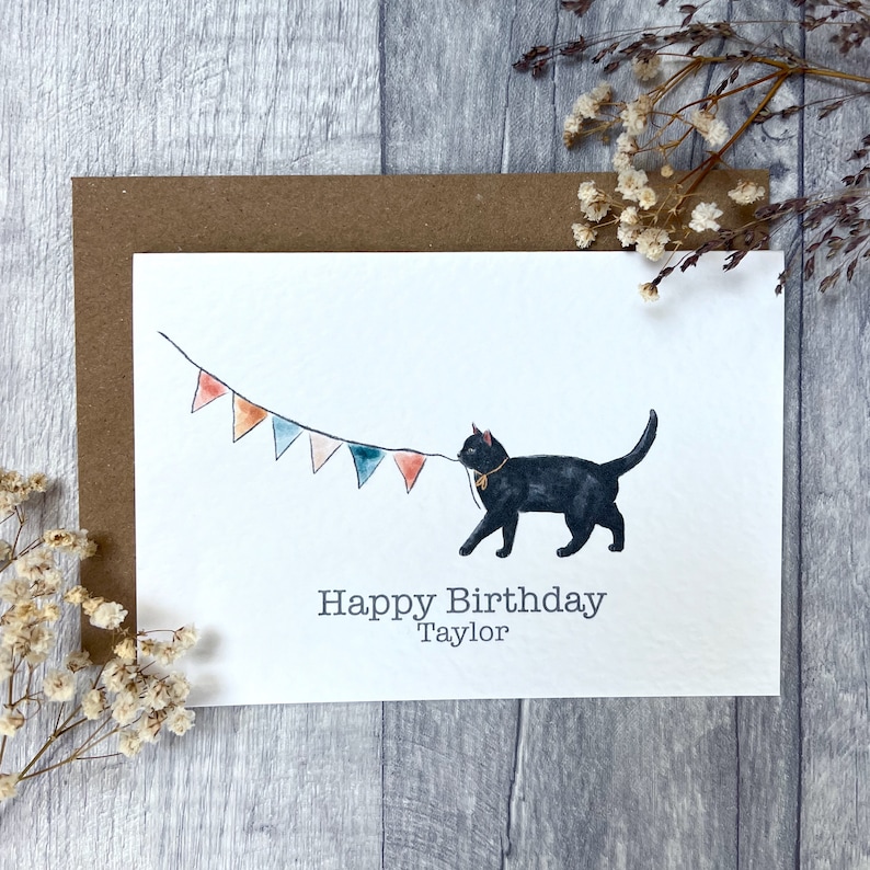 May include: A white greeting card featuring a watercolor illustration of a black cat holding a string of colorful triangular bunting flags in its mouth. Below the cat, the text reads Happy Birthday Taylor in a simple gray sans-serif font.