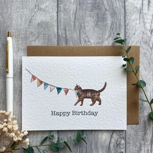 May include: A white greeting card featuring a watercolor illustration of a brown tabby cat walking and holding a string of colorful triangular bunting flags in its mouth. The text Happy Birthday is printed in black below the cat illustration.