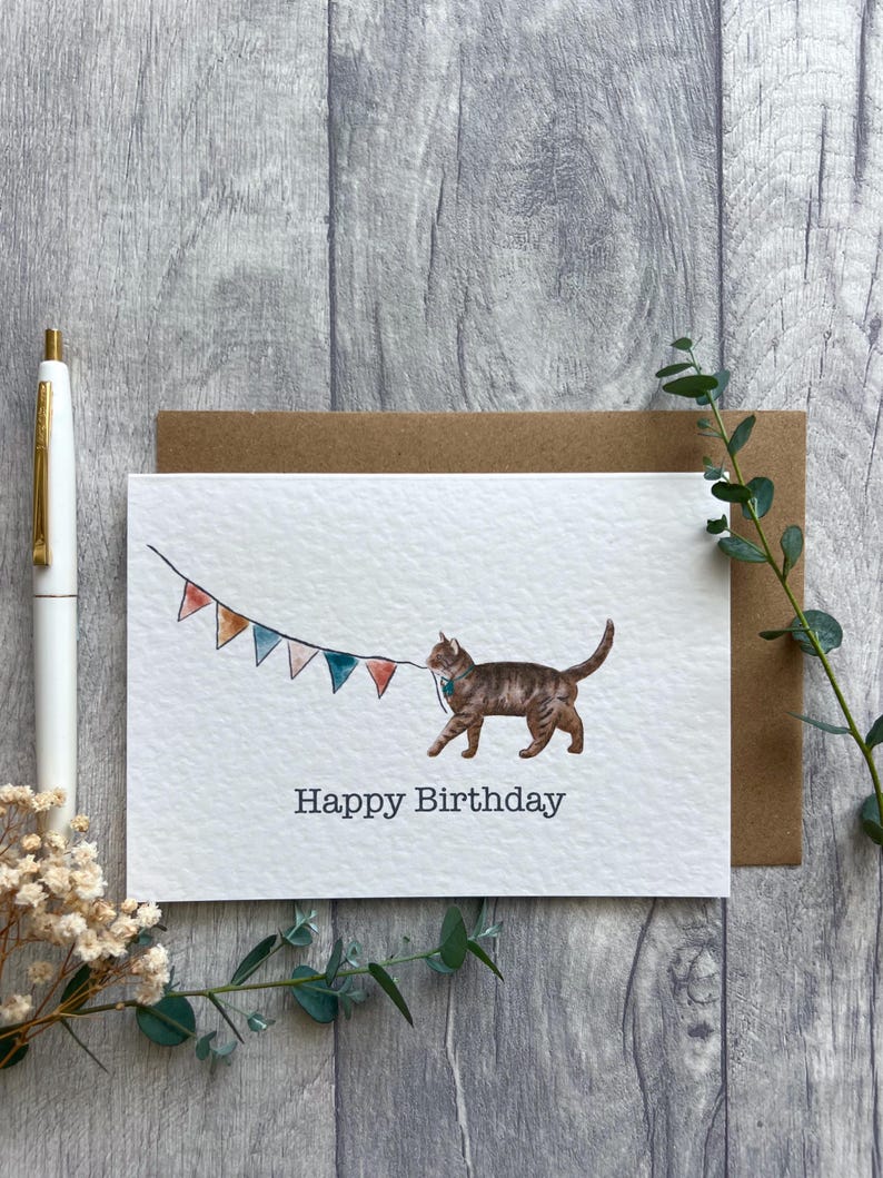 May include: A white greeting card featuring a watercolor illustration of a brown tabby cat walking and holding a string of colorful triangular bunting flags in its mouth. The text Happy Birthday is printed in black below the cat illustration.