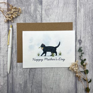May include: A greeting card with a black cat carrying flowers, with the text "Happy Mother's Day". The card is on a wooden surface, with a pen and envelope nearby. The cat illustration is in a watercolor style.