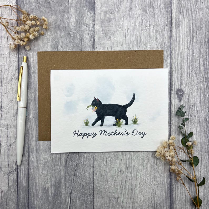 May include: A greeting card with a black cat carrying flowers, with the text "Happy Mother's Day". The card is on a wooden surface, with a pen and envelope nearby. The cat illustration is in a watercolor style.
