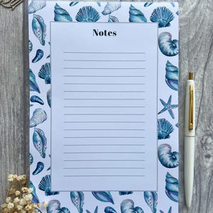 May include: A white notepad with a blue and white seashell pattern border. The notepad has lined paper and the word "Notes" printed at the top. A white pen is lying next to the notepad.