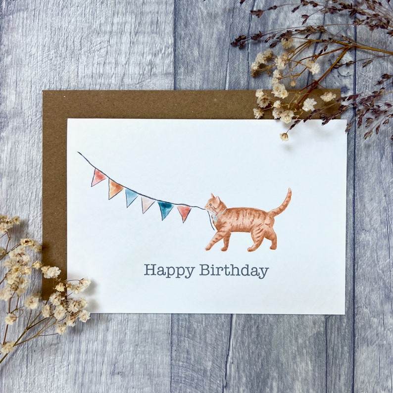 May include: A white card featuring a watercolour illustration of an orange tabby cat pulling a string of colourful bunting. The text "Happy Birthday" is printed below the cat.