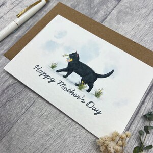May include: Greeting card with a black cat carrying flowers, with the text "Happy Mother's Day." The card is on a brown envelope, with a white pen in the background. The background is a light gray wood.