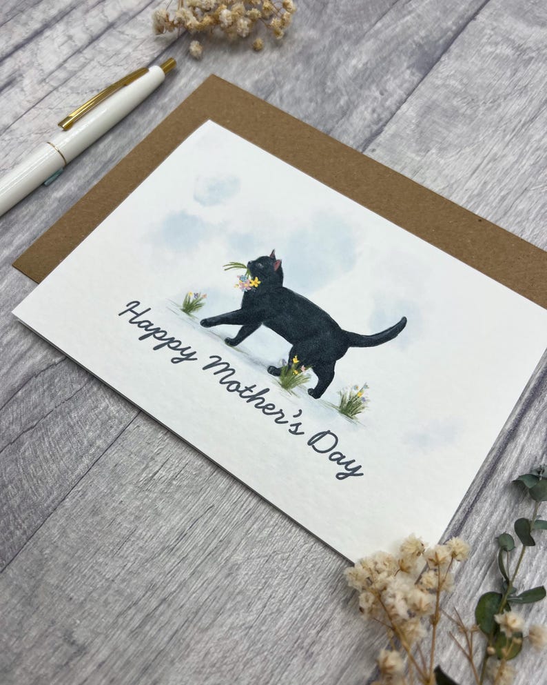 May include: Greeting card with a black cat carrying flowers, with the text "Happy Mother's Day." The card is on a brown envelope, with a white pen in the background. The background is a light gray wood.