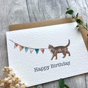 May include: A white greeting card featuring a watercolor illustration of a brown tabby cat holding a string of colorful triangular bunting flags in its mouth. The text Happy Birthday is printed in a dark gray sans-serif font below the cat.