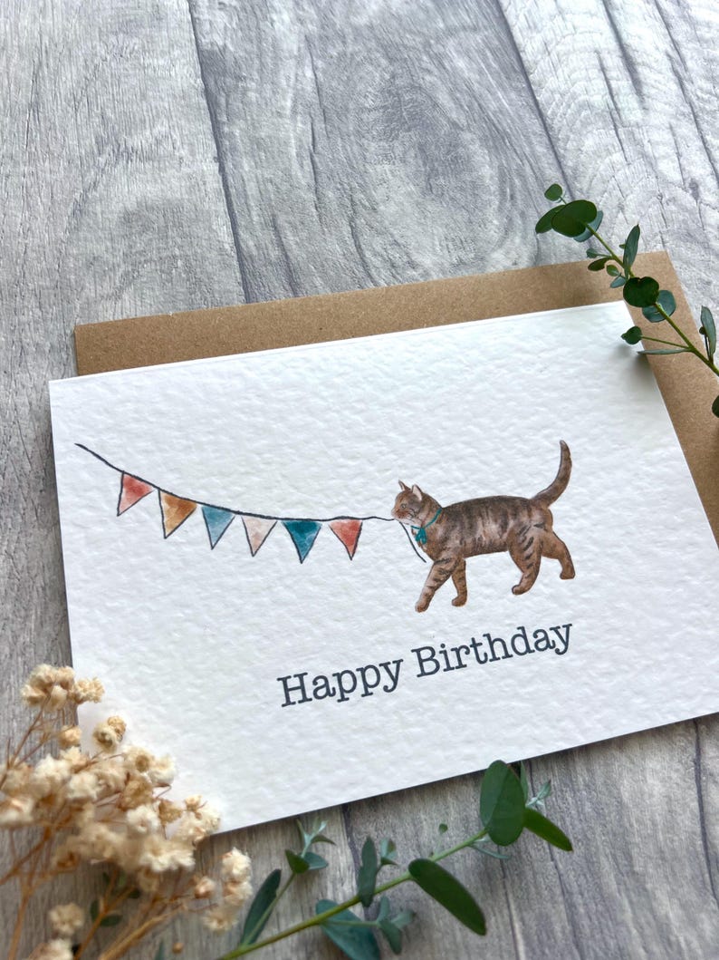 May include: A white greeting card featuring a watercolor illustration of a brown tabby cat holding a string of colorful triangular bunting flags in its mouth. The text Happy Birthday is printed in a dark gray sans-serif font below the cat.