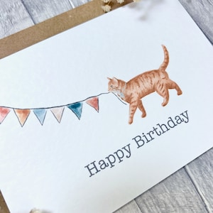 May include: A white card featuring a watercolour illustration of an orange cat pulling a string of colourful bunting. The text "Happy Birthday" is printed below the cat.