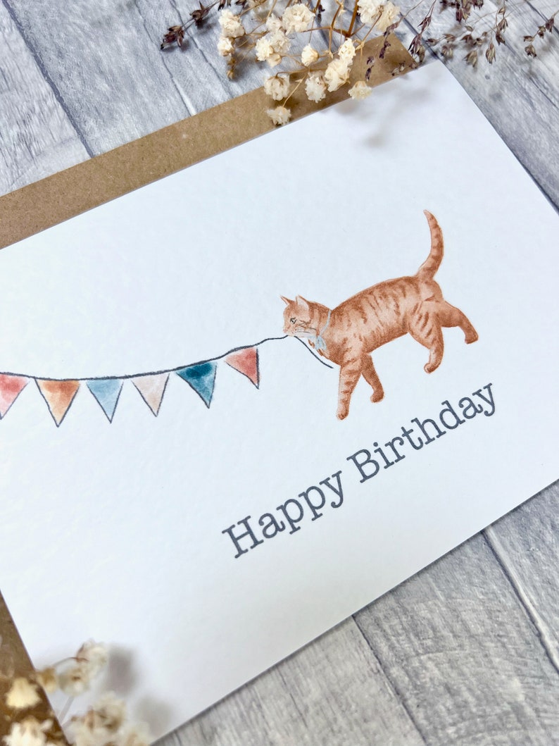 May include: A white card featuring a watercolour illustration of an orange cat pulling a string of colourful bunting. The text "Happy Birthday" is printed below the cat.