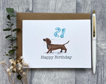 Puede incluir: Una tarjeta de felicitación blanca con la ilustración de un perro salchicha marrón que sostiene globos azules con forma del número 21 y el texto Happy Birthday impreso en gris oscuro. La tarjeta está sobre un sobre marrón junto a un bolígrafo blanco.