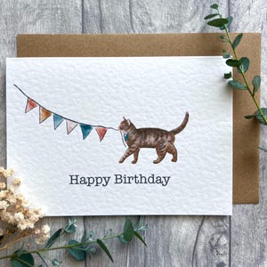 May include: A white greeting card featuring a watercolor illustration of a brown tabby cat wearing a teal collar and pulling a string of colorful triangular bunting flags. The text Happy Birthday is printed in dark gray below the cat.