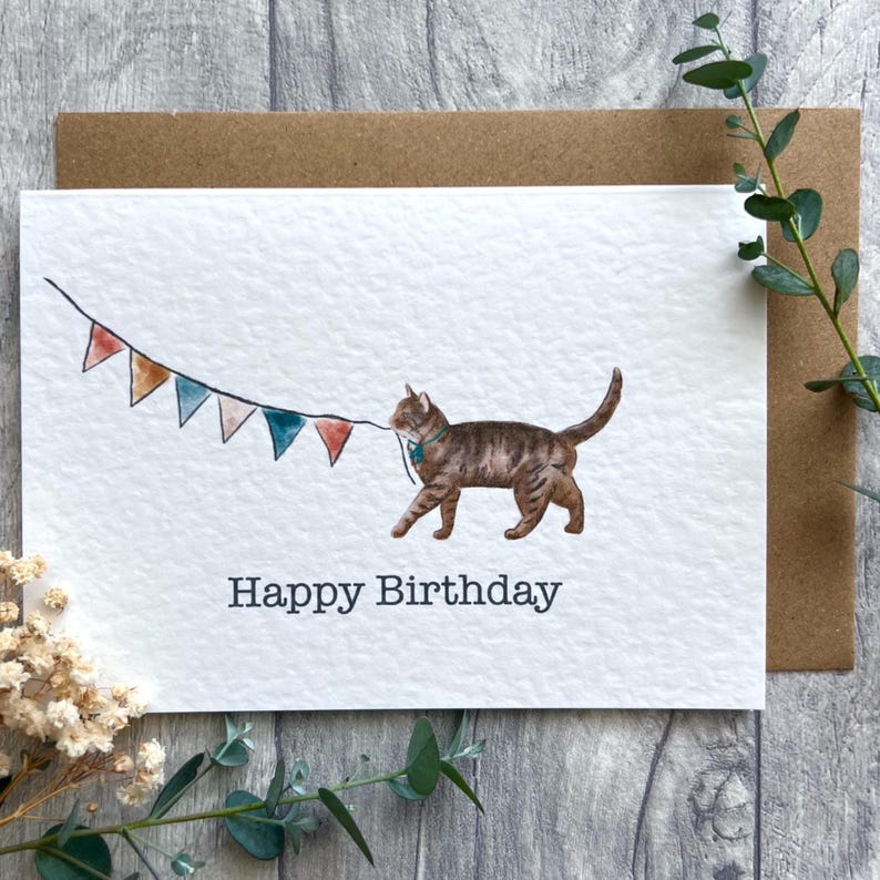 May include: A white greeting card featuring a watercolor illustration of a brown tabby cat wearing a teal collar and pulling a string of colorful triangular bunting flags. The text Happy Birthday is printed in dark gray below the cat.