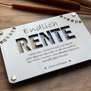 Könnte beinhalten: Eine Holzkiste mit einem Ausschnitt des Wortes "RENTE" und einer Nachricht, die jemandem eine glückliche Rente wünscht. Die Kiste ist mit kleinen Flaggen verziert und hat einen schwarzen Rand.