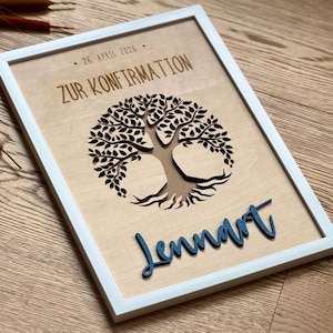 May include: A white framed wall art piece featuring a light wood background with a laser-cut tree silhouette. The text reads "ZUR KONFIRMATION", "26. APRIL 2026", and the name "Lennart" in raised, dark blue script lettering at the bottom.