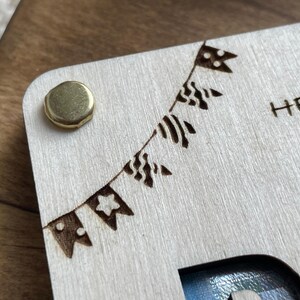 May include: A wooden box with a cutout for a photo and a brass hinge. The box has a decorative design of a string of flags with a star, polka dots, and stripes. The text "HERZ" is engraved on the box.