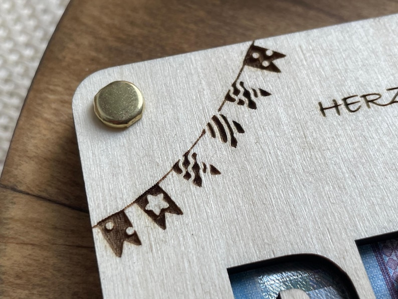 May include: A wooden box with a cutout for a photo and a brass hinge. The box has a decorative design of a string of flags with a star, polka dots, and stripes. The text "HERZ" is engraved on the box.