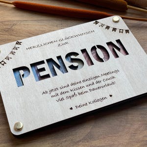 May include: A wooden sign with the word "PENSION" cut out and the text "HERZLICHEN GL&Uuml;CKWUNSCH ZUR PENSION Ab jetzt sind deine einzigen Meetings mit dem Kissen und der Couch. Viel Spa&szlig; beim Dauerurlaub! Deine Kollegen" written on it.