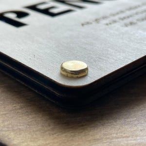 May include: Close-up of a wooden sign with the word "PEN" in black lettering. The sign is attached to a wooden surface with a gold metal screw.
