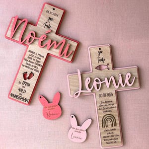 Könnte beinhalten: Zwei Holzkreuze mit rosa Akzenten und den Namen "Noemi" und "Leonie" werden angezeigt. Jedes Kreuz hat ein Datum und Text. Zwei rosa hasenförmige Ornamente sind ebenfalls vorhanden, eines mit dem Namen "Noemi" und das andere mit "Leonie".