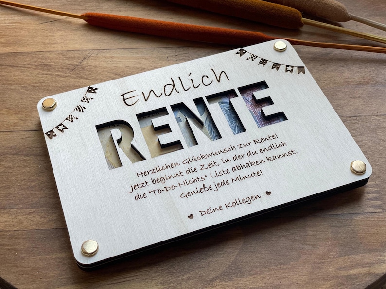 Könnte beinhalten: Eine Holzkiste mit einem Ausschnitt des Wortes "RENTE" und einer Nachricht, die jemandem eine glückliche Rente wünscht. Die Kiste ist mit kleinen Flaggen verziert und hat einen schwarzen Rand.