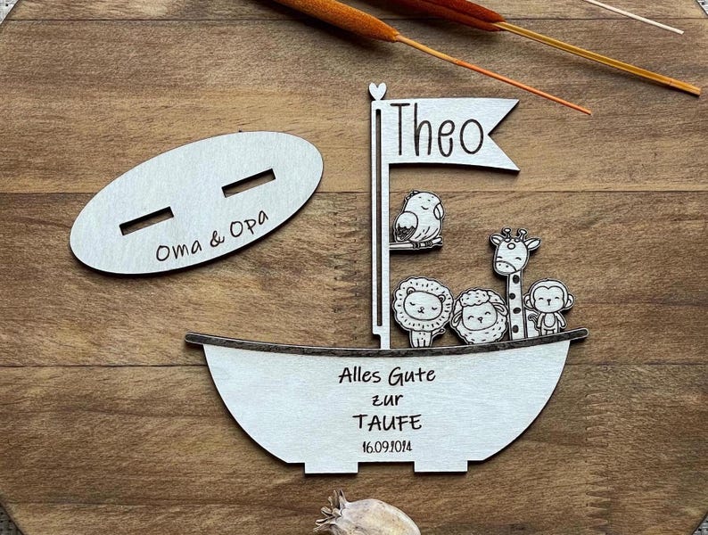 May include: Wooden boat-shaped sign with the name "Theo" on the sail, a giraffe, lion, monkey, and bird, and the text "Alles Gute zur Taufe 16.09.2014". There is a separate oval-shaped sign with the text "Oma & Opa".